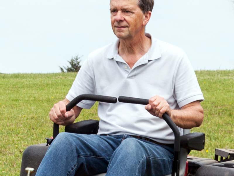 An elderly man mowing a large field of grass with a zero turn mower.